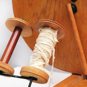 May include: A wooden yarn winder with two spools. One spool is empty, the other is filled with cream-coloured yarn. The winder is made of light and dark brown wood and has a black metal support.