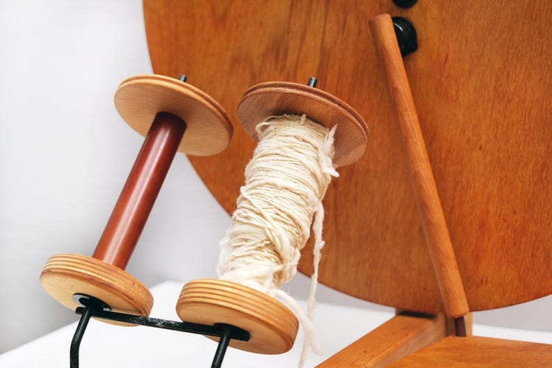 May include: A wooden yarn winder with two spools. One spool is empty, the other is filled with cream-coloured yarn. The winder is made of light and dark brown wood and has a black metal support.