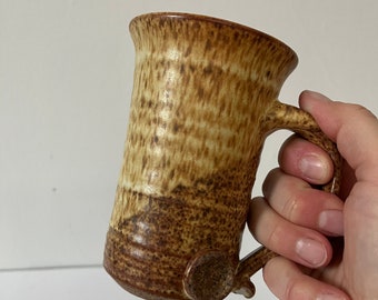 May include: A brown and tan ceramic mug with a speckled glaze. The mug has a rounded handle and a slightly flared rim.