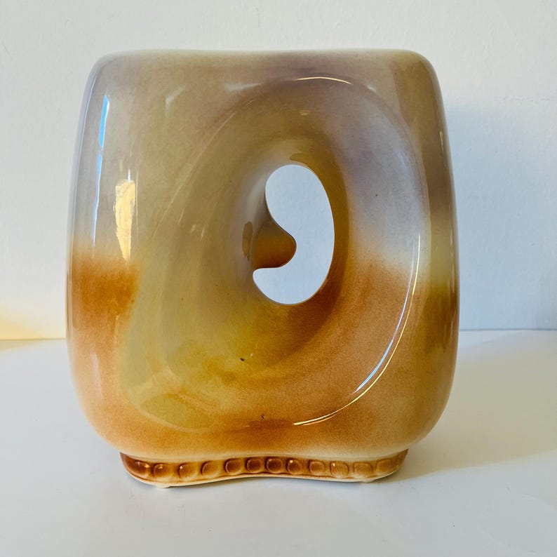 Może przedstawiać: Ceramiczny wazon o unikalnym designie. Wazon ma kwadratowy kształt z dużym, zakrzywionym otworem pośrodku. Kolory przechodzą od jasnobrązowego do ciemnobrązowego, z błyszczącym wykończeniem. Podstawa ma dekoracyjny, teksturowany wz&oacute;r.