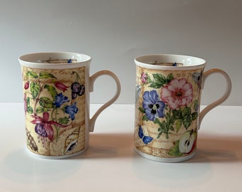 May include: Two white ceramic mugs with floral designs. The mugs have a cream background with a floral pattern in shades of pink, blue, purple, and green. The mugs have a handwritten text pattern on the background.