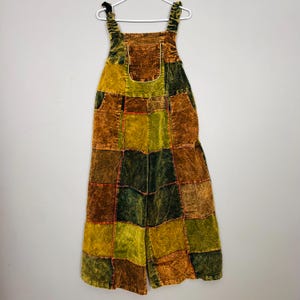 Può includere: Una tuta patchwork nei toni del marrone, verde e giallo è appesa a una gruccia bianca. La tuta presenta un davanti a pettorina, spalline regolabili e gambe larghe. Il tessuto sembra essere velluto a coste o un materiale strutturato simile, con cuciture visibili.