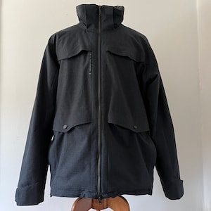 May include: A black, zippered jacket with a high collar and two large pockets. The jacket has a textured fabric and a subtle sheen.