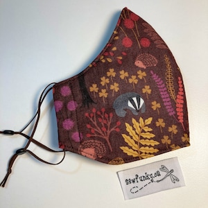 May include: A brown fabric face mask with a woodland pattern featuring hedgehogs, badgers, and leaves. The mask has brown ties and a white tag with the text "sewfunky.ca" and a dragonfly.