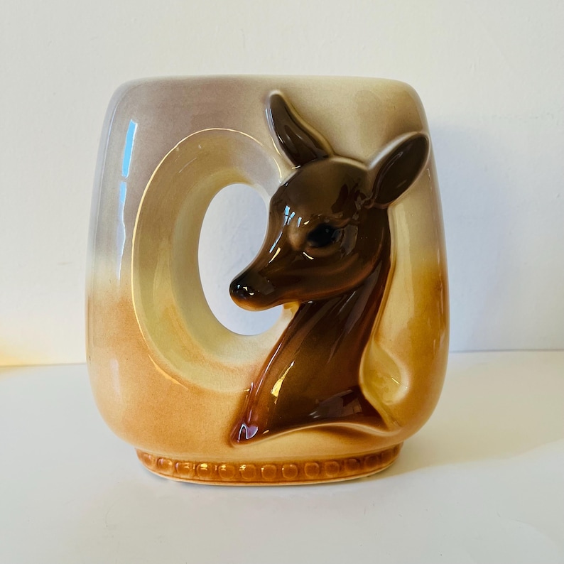 Może przedstawiać: Ceramiczny wazon z motywem głowy jelenia. Wazon ma prostokątny kształt z dużym okrągłym wycięciem. Głowa jelenia jest brązowa, a wazon ma gradient kolor&oacute;w beżowego i brązowego. Podstawa ma wz&oacute;r wytłoczony.