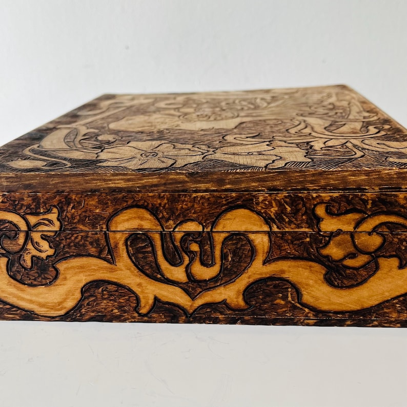May include: A wooden box with intricate carvings. The box features a dark brown exterior with lighter brown carved floral and scroll designs. The top and sides are adorned with detailed patterns, creating a decorative storage piece.