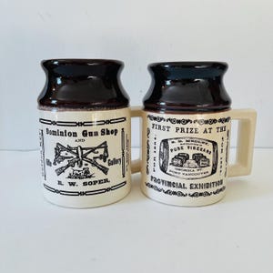 Puede incluir: Dos tazas de cerámica marrón y blanca con letras negras. La taza de la izquierda tiene el texto "Dominion Gun Shop and Gallery R. W. Soper." La taza de la derecha tiene el texto "First Prize at the Provincial Exhibition for 1804 S. B. Medley, Manufacturer of Pure Vinegars, Georgia St. Port Vancouver."