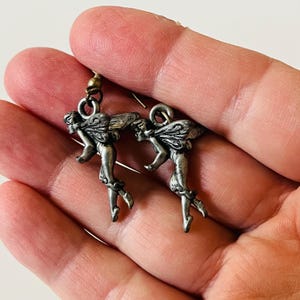 May include: A pair of silver-coloured fairy earrings. Each earring features a detailed fairy figure with outstretched wings, suspended from a small hook. The fairies are in a flying pose, with intricate detailing on their wings and bodies. The earrings are held in a hand.
