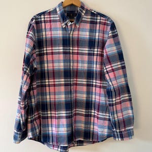 Peut inclure: Une chemise à carreaux à manches longues dans les tons de rose, de bleu et de marine. La chemise a un col boutonné et un petit logo brodé sur la poitrine. La chemise est suspendue à un cintre en bois.