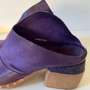 May include: A pair of purple leather ankle boots with a stacked heel. The boots have a textured design on the heel and are embellished with small gold-colored studs. The words "MADE IN ITALY" are visible inside the boot.