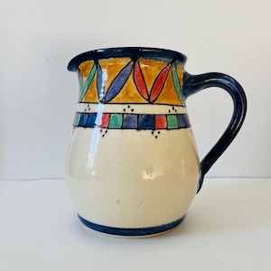 Peut inclure: Une cruche en céramique avec un corps crème et une anse et un bord bleu foncé. La cruche est décorée de motifs géométriques colorés en jaune, rouge, bleu et vert. Le design comprend une bande de carrés et une série de formes en forme de feuilles.