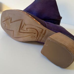 May include: A close-up of a purple suede boot with a wooden heel. The sole is brown with a star and arrow design. The heel is approximately 2 inches high. The size is marked as 37.