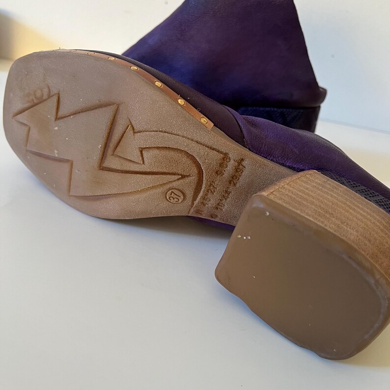 May include: A close-up of a purple suede boot with a wooden heel. The sole is brown with a star and arrow design. The heel is approximately 2 inches high. The size is marked as 37.