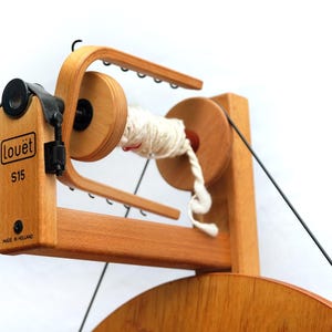 May include: A wooden Lou&euml;t S15 spinning wheel, featuring a light wood finish. The wheel has a spindle with white yarn, and the text "Lou&euml;t S15 Made in Holland". This spinning wheel is designed for yarn and fibre craft projects.