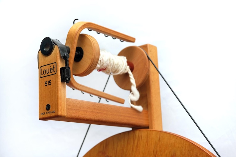 May include: A wooden Lou&euml;t S15 spinning wheel, featuring a light wood finish. The wheel has a spindle with white yarn, and the text "Lou&euml;t S15 Made in Holland". This spinning wheel is designed for yarn and fibre craft projects.