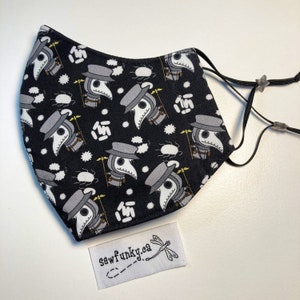 Puede incluir: Mascarilla negra con un patrón repetido de ilustraciones de médicos de la peste en gris, blanco y beige. La mascarilla tiene presillas ajustables negras y una pequeña etiqueta blanca con el texto "sewfunky.ca" y un logotipo de libélula.