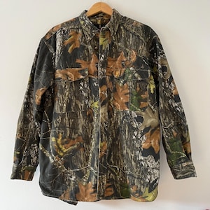 May include: A long-sleeved button-up shirt in a camouflage pattern with brown, green, and black colors. The shirt has two flap pockets on the front.