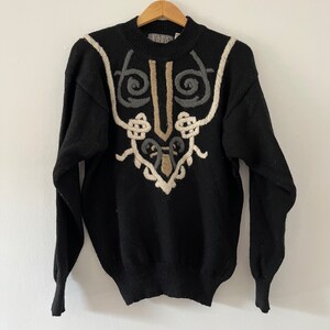 May include: Black sweater with a unique embroidered design in beige, gray, and off-white. The sweater features a crew neck and long sleeves. The embroidered pattern is symmetrical and abstract, adding a vintage touch to the garment.