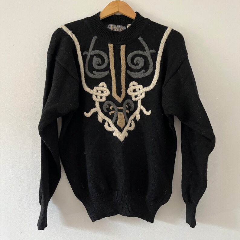 May include: Black sweater with a unique embroidered design in beige, gray, and off-white. The sweater features a crew neck and long sleeves. The embroidered pattern is symmetrical and abstract, adding a vintage touch to the garment.