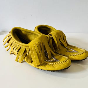 May include: A pair of yellow leather moccasins with fringe detailing. The shoes have a rounded toe and are decorated with a beaded design on the vamp. The soles are dark brown.