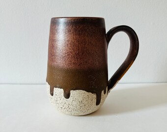 May include: A brown and white ceramic mug with a speckled glaze. The mug has a rounded body and a handle.