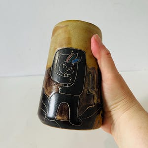 May include: A ceramic vase with a tan and brown gradient. The vase features a black illustration of a figure with a rabbit head and a cactus. The figure is hugging itself. The vase is held by a hand.