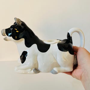 May include: A ceramic cow creamer in a sitting position, with a black and white spotted pattern. The creamer has a curved handle and a spout on its back. The cow's horns and hooves are gray.