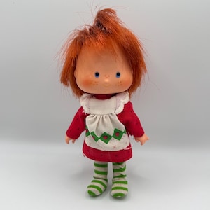 May include: A vintage doll with red hair, blue eyes, and freckles. The doll wears a red dress with a white apron featuring green heart and diamond patterns, and green and white striped socks. A collectible toy.