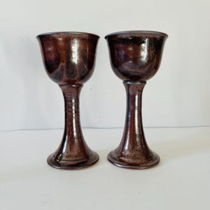 Könnte beinhalten: Zwei braune Keramikkelche mit einem gesprenkelten Überzug. Die Kelche haben eine breite Schale und einen hohen, schlanken Stiel.