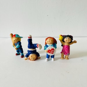 Op de afbeelding: Vier Cabbage Patch Kids-figuurtjes. Eén draagt een honkbaluniform en houdt een knuppel vast, een ander staat op zijn handen, een ander houdt een hartvormig bord vast met de tekst "Ik hou van je", en de laatste draagt een roze en witte badpak en houdt een gele rubberen eend vast.