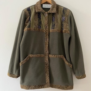 May include: Olive green fleece jacket with brown patterned accents. The jacket features a button-up front, two pockets, and decorative trim around the collar, pockets, and edges. The upper portion has a patchwork design with floral and geometric patterns.