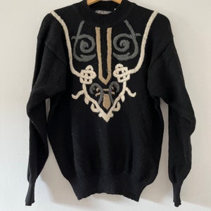 May include: Black sweater with a crew neck and long sleeves. The sweater features an embroidered design in cream, gray, and tan colors on the front. The design has a symmetrical, ornate pattern.
