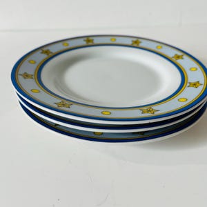 Könnte beinhalten: Ein Stapel aus drei weißen Keramiktellern mit einem breiten hellblauen Rand, der mit einem wiederkehrenden Muster aus gelben Sternen und kleinen gelben Kreisen verziert ist, umrandet von dünnen dunkelblauen und gelben Ringen.