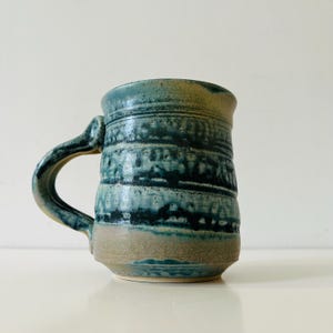 May include: A ceramic mug featuring a textured, mottled glaze in shades of deep teal, navy blue, and sandy beige. The vessel has a cylindrical body with a prominent, thick handle attached to the side, showcasing a layered, reactive finish.