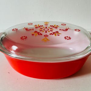 May include: A vintage red Pyrex casserole dish with a clear glass lid. The lid features a floral and bird pattern in red, yellow, and orange. The dish is round with a bright red exterior.