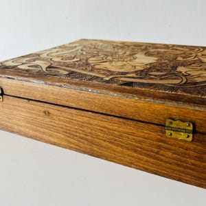 May include: A wooden box with a detailed carved design on the lid. The box is stained a warm brown color and has brass hinges. The carved design appears to be a floral or mythological theme.