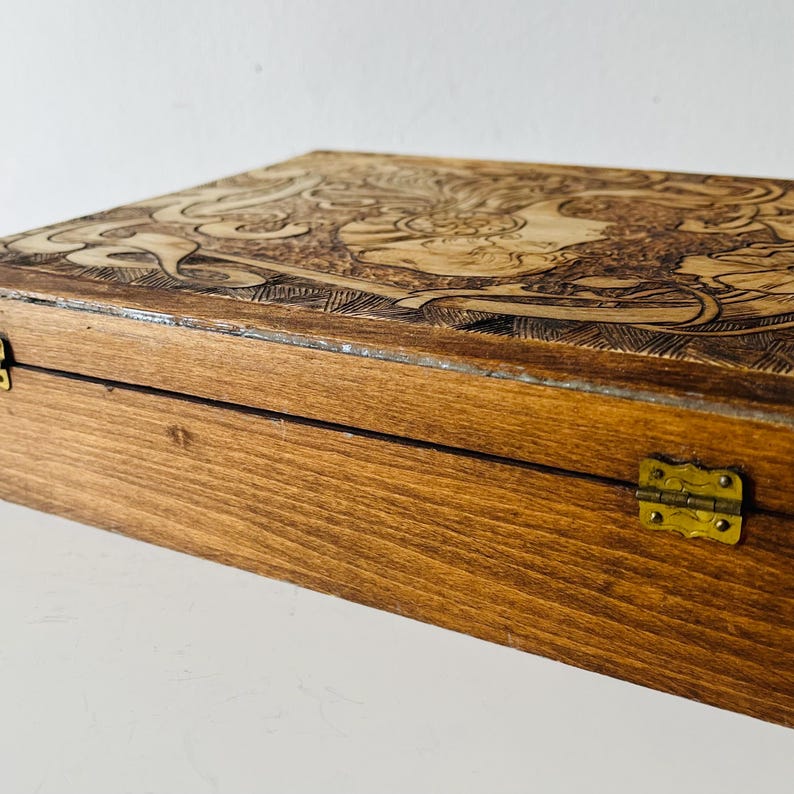 May include: A wooden box with a detailed carved design on the lid. The box is stained a warm brown color and has brass hinges. The carved design appears to be a floral or mythological theme.