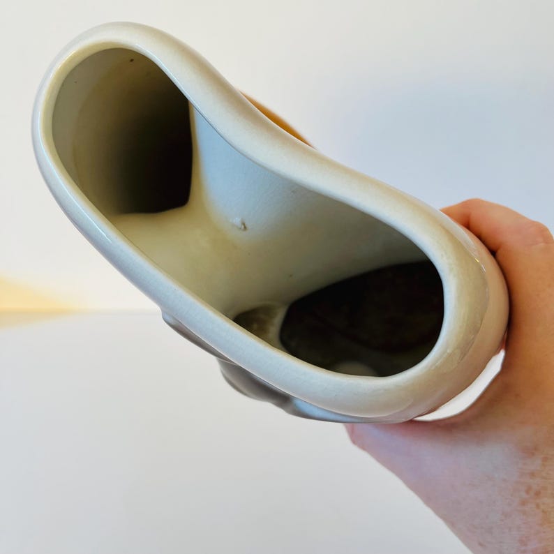 Może przedstawiać: Kremowy ceramiczny wazon o unikalnym, asymetrycznym kształcie. Wazon ma szeroki, otwarty wierzch i wąską podstawę. Wnętrze jest ciemnobrązowe, kontrastujące z zewnętrzem. Wazon jest trzymany przez dłoń.