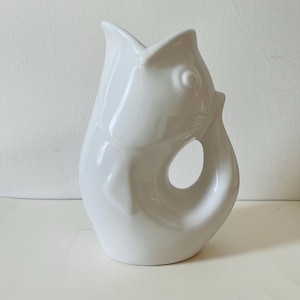 May include: A white ceramic fish-shaped jug. The jug features a pouring spout, a handle formed by the fish's tail, and a curved opening on the side. The design includes a fish eye. The jug is suitable for serving drinks.