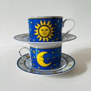 Könnte beinhalten: Zwei gestapelte Keramiktassen mit Untertassen, die einen leuchtend blauen Hintergrund mit gelben Himmelsmotiven aufweisen. Die obere Tasse zeigt eine Sonne mit Gesicht, während die untere Tasse einen Halbmond und gelbe Sterne als Dekor trägt.