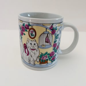 May include: A ceramic mug with a decorative design featuring a white cat with a pink bow, a bird in a cage, and pink roses. The mug has a light blue handle and rim, with a yellow background.