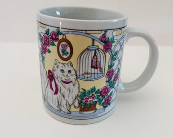 May include: A ceramic mug with a decorative design featuring a white cat with a pink bow, a bird in a cage, and pink roses. The mug has a light blue handle and rim, with a yellow background.