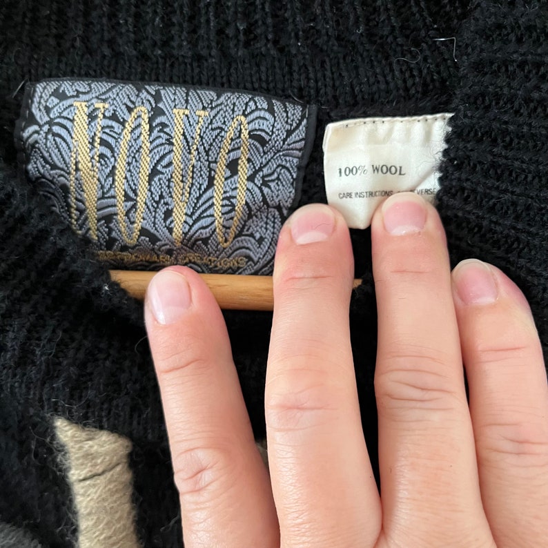 May include: Close-up of a black wool sweater with a tag that reads "100% Wool". The sweater has a textured knit pattern. A second tag features the brand name in gold lettering on a patterned background.