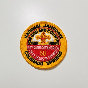 Puede incluir: Un parche circular con borde amarillo y centro rojo y blanco. El parche dice "NATIONAL JAMBOREE FOR GOD AND COUNTRY" y "BOY SCOUTS OF AMERICA". Conmemora "FIFTY YEARS OF SERVICE" en Colorado Springs.
