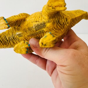 Puede incluir: Un juguete de dinosaurio Apatosaurus de pl&aacute;stico amarillo con detalles azules en los pies. La parte inferior del juguete es visible, con el texto "&copy; 1991 U.K.R.D. NO. 2022243 APATOSAURUS" y "MADE IN CHINA" impreso. El juguete est&aacute; sostenido en una mano.