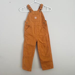 Peut inclure: Une salopette orange-brun pour enfant suspendue à un cintre blanc. La salopette a des bretelles réglables, une poche de bavette et deux poches avant. Les genoux ont des patchs renforcés. Un petit logo carré est sur la poche de la bavette.