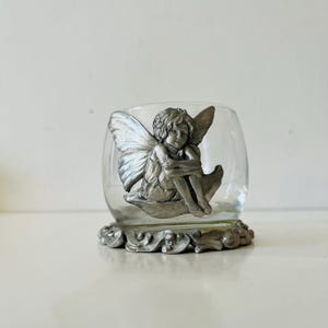 May include: A clear glass votive candle holder with a decorative silver-colored fairy figurine. The fairy has detailed wings and is seated on a leaf. The base of the holder has an ornate, scrolling design.