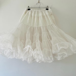 Può includere: Una sottogonna bianca con più strati di tulle e una fascia in vita arricciata. La gonna è appesa a un gancio contro un muro bianco. La gonna è progettata per aggiungere volume a un vestito o una gonna.