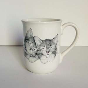 May include: White ceramic mug featuring a charming illustration of two gray and white kittens snuggled together. The kittens have their eyes closed and pink noses. The mug has a curved handle and a simple, elegant design, perfect for cat lovers.
