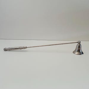 May include: A silver-toned candle snuffer with a bell-shaped head and a long, slender handle. The handle has a decorative, textured grip. The snuffer is designed to extinguish candles safely and efficiently.